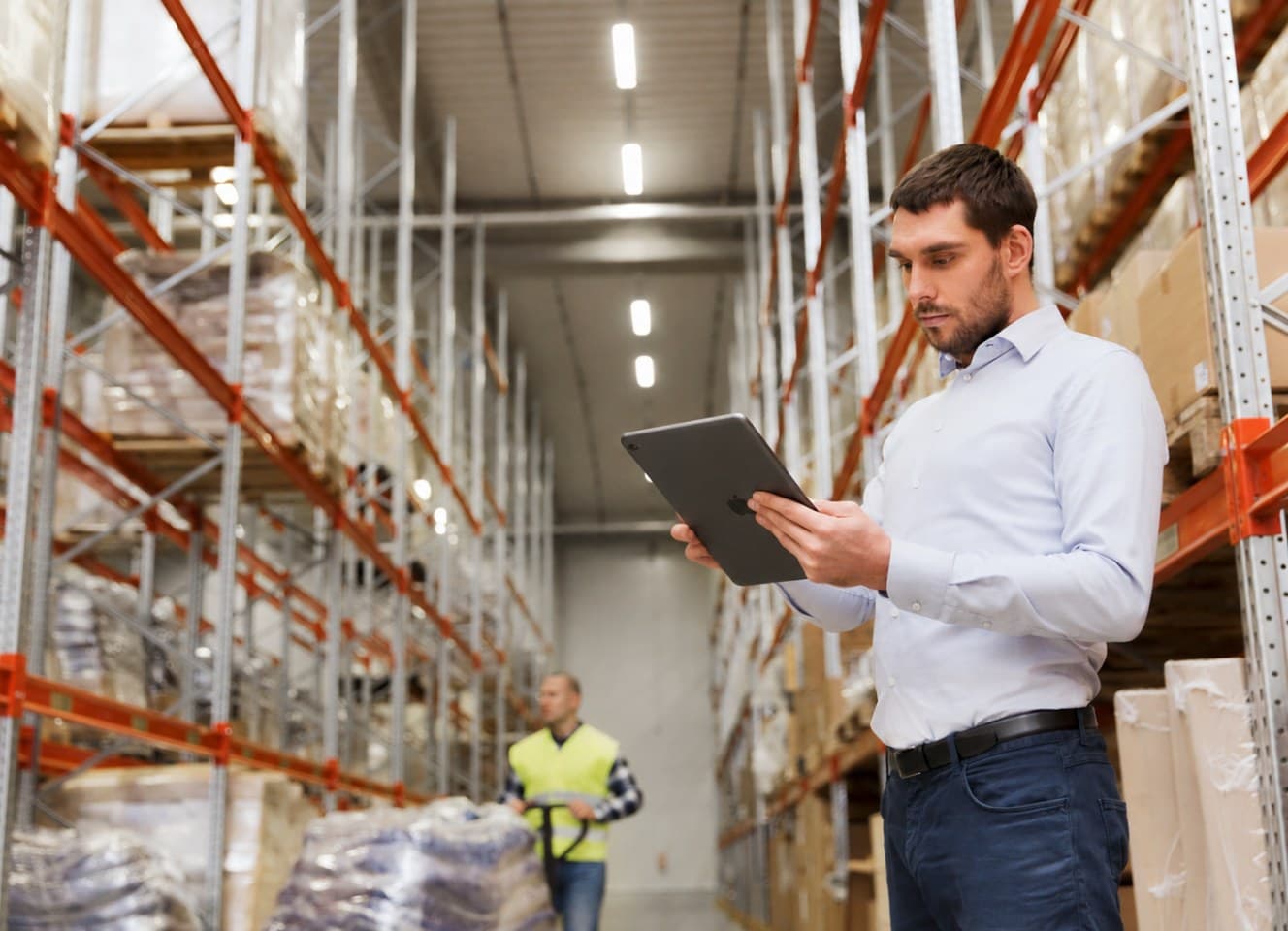 A man holding a tablet in a warehouse aisle between tall pallet racks, with a colleague in a hi-vis vest working further down the aisle