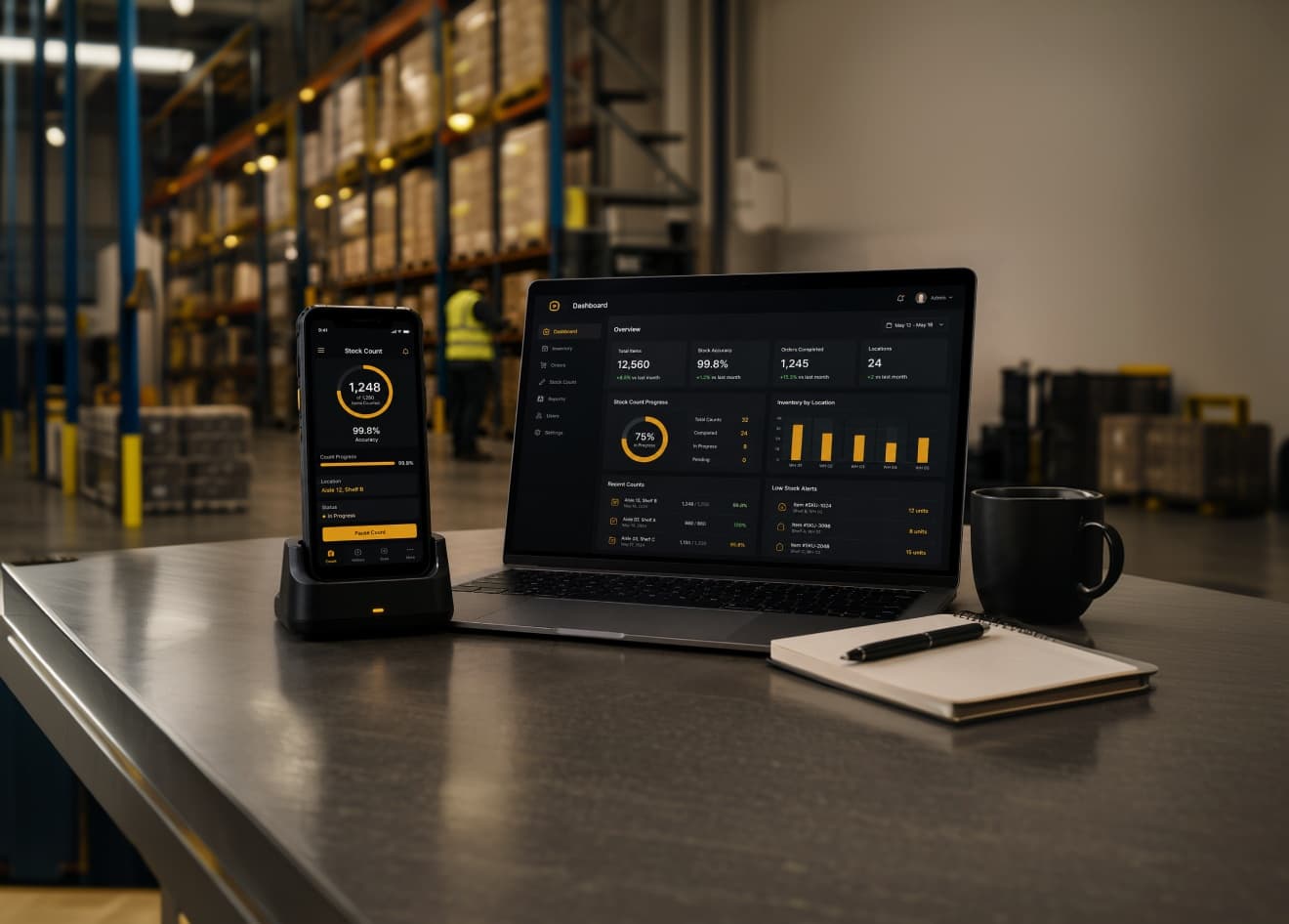 A rugged handheld in a charging dock showing a stock count at 98.6 per cent, beside a laptop showing the Footprints web dashboard, on a warehouse packing table with a notebook and a mug